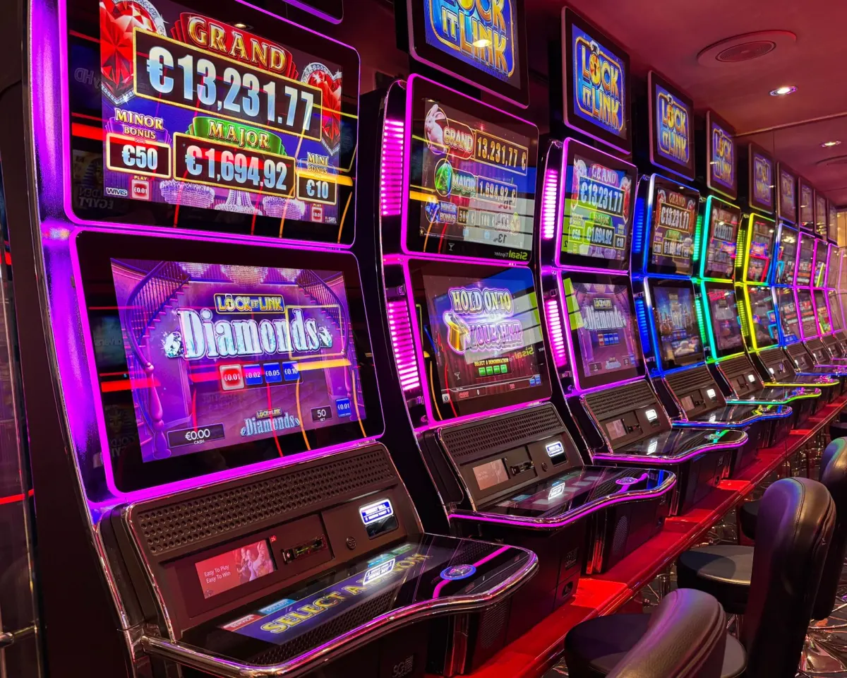 A row of brightly lit casino slot machines with jackpot displays on their top screens and empty stools in front